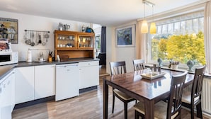 Une cuisine moderne avec des meubles blancs, une table à manger en bois et une vue sur les arbres par une grande fenêtre.