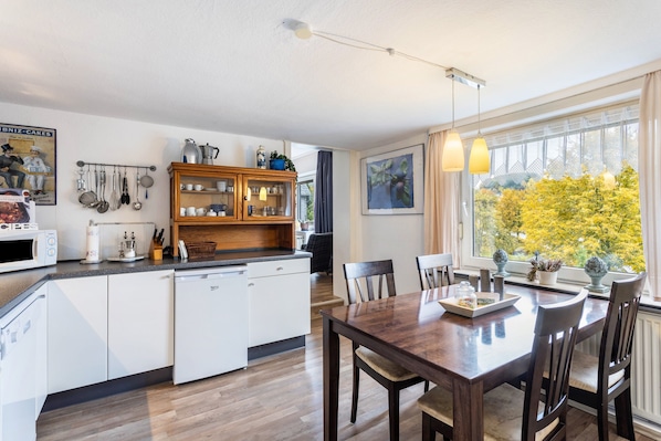 A modern kitchen with white cabinetry, a wooden dining table, and a view of trees through a large window.