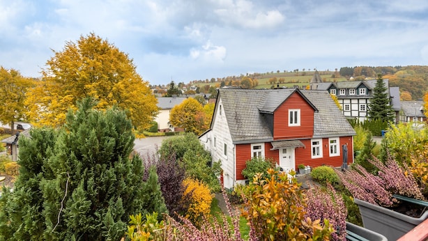 Une maison rouge avec une porte blanche, entourée d’un feuillage automnal et sous un ciel nuageux.