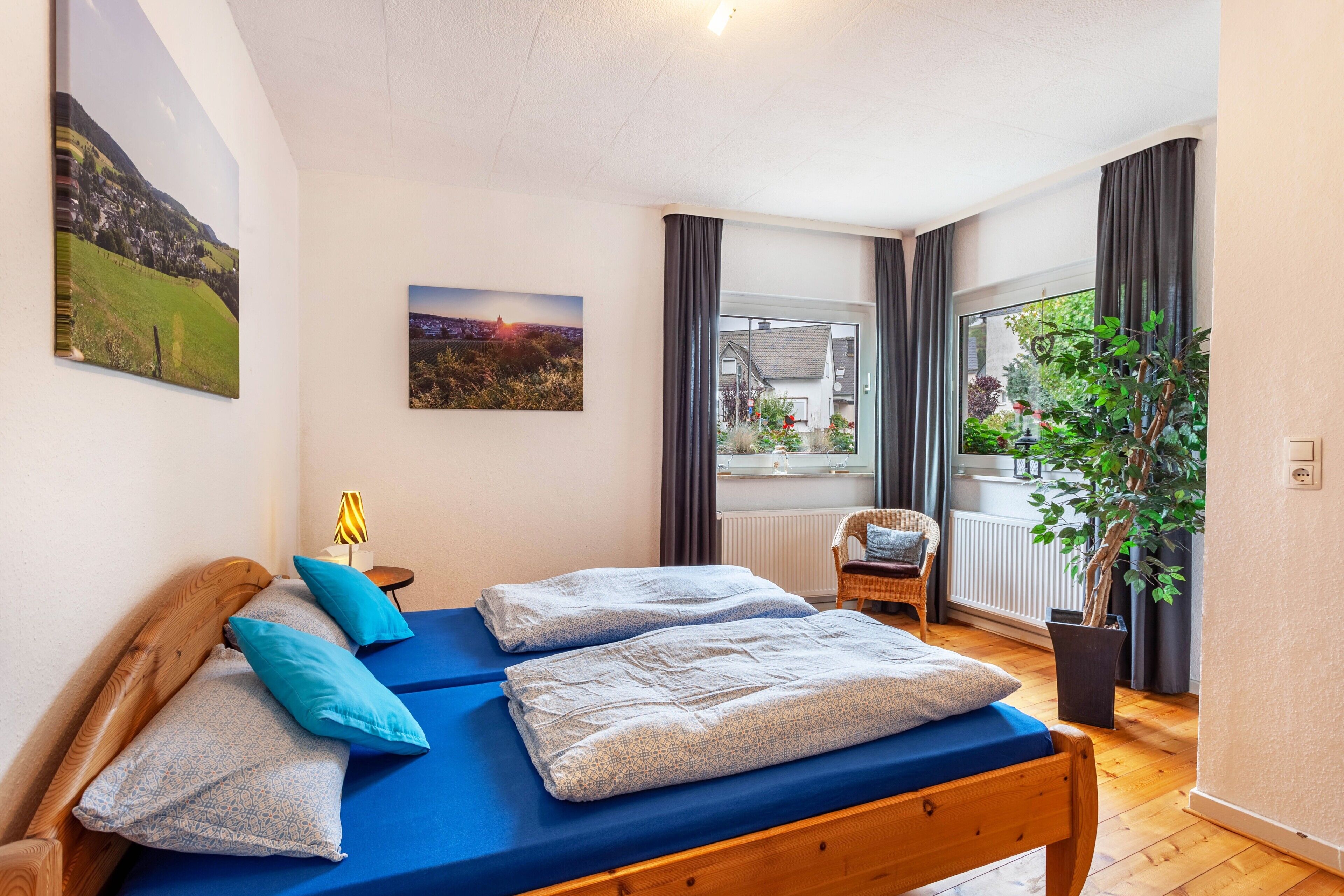 A bedroom with a wooden bed, two pillows, a blue mattress, a window with curtains, a potted plant, and a view of a building outside.