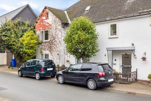 Een woonstraat met twee geparkeerde auto's, een wit huis met een rood dak en een boom voor het huis.