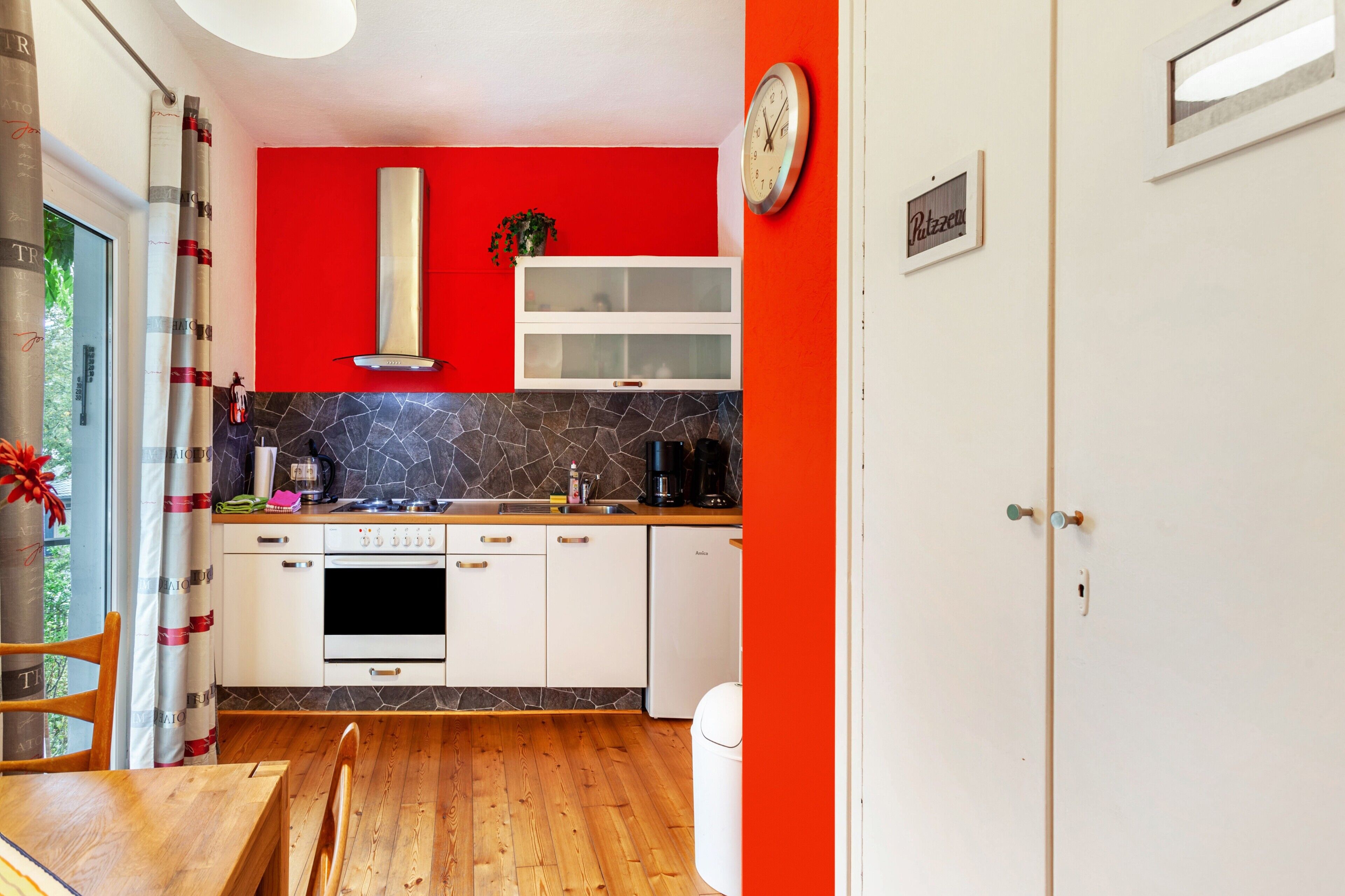 A kitchen with white cabinets, a stone backsplash, a red accent wall, a clock, and a nameplate.