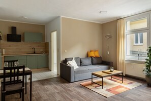 A compact living space with a kitchenette, a sofa with cushions, a dining table with chairs, and a large window with curtains. - Apartment 'Innenstadt Quartier' (Neuruppin)