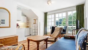 Un vaste espace de vie comprenant un canapé, une table en bois et des chaises, une cuisine à l’arrière-plan et une grande fenêtre avec des rideaux.