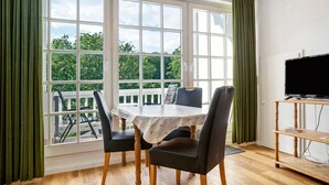Une pièce avec une table et des chaises, un téléviseur posé sur un meuble, et une grande fenêtre avec des rideaux.
