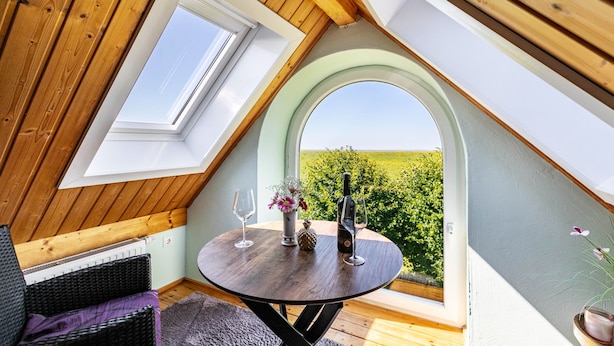 Ein Zimmer mit schräg verlaufender Decke, einem runden Tisch mit Flasche und Glas, und Blick ins Grüne.