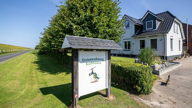 A sign for a property named 'Ferienwohnung Kiebitznest' with a house in the background. - Apartment 'Kiebeitznest' with Sea View, Private Terrace and Wi-Fi (Kaiser-Wilhelm-Koog)