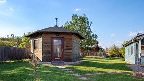 Una pequeña cabaña de madera con una puerta roja, rodeada por un césped cuidado y una valla de madera.