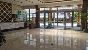 A hotel lobby with a marble reception desk, glass doors leading outside, and a large potted plant. - Baibuting Hotel (Haiyan)