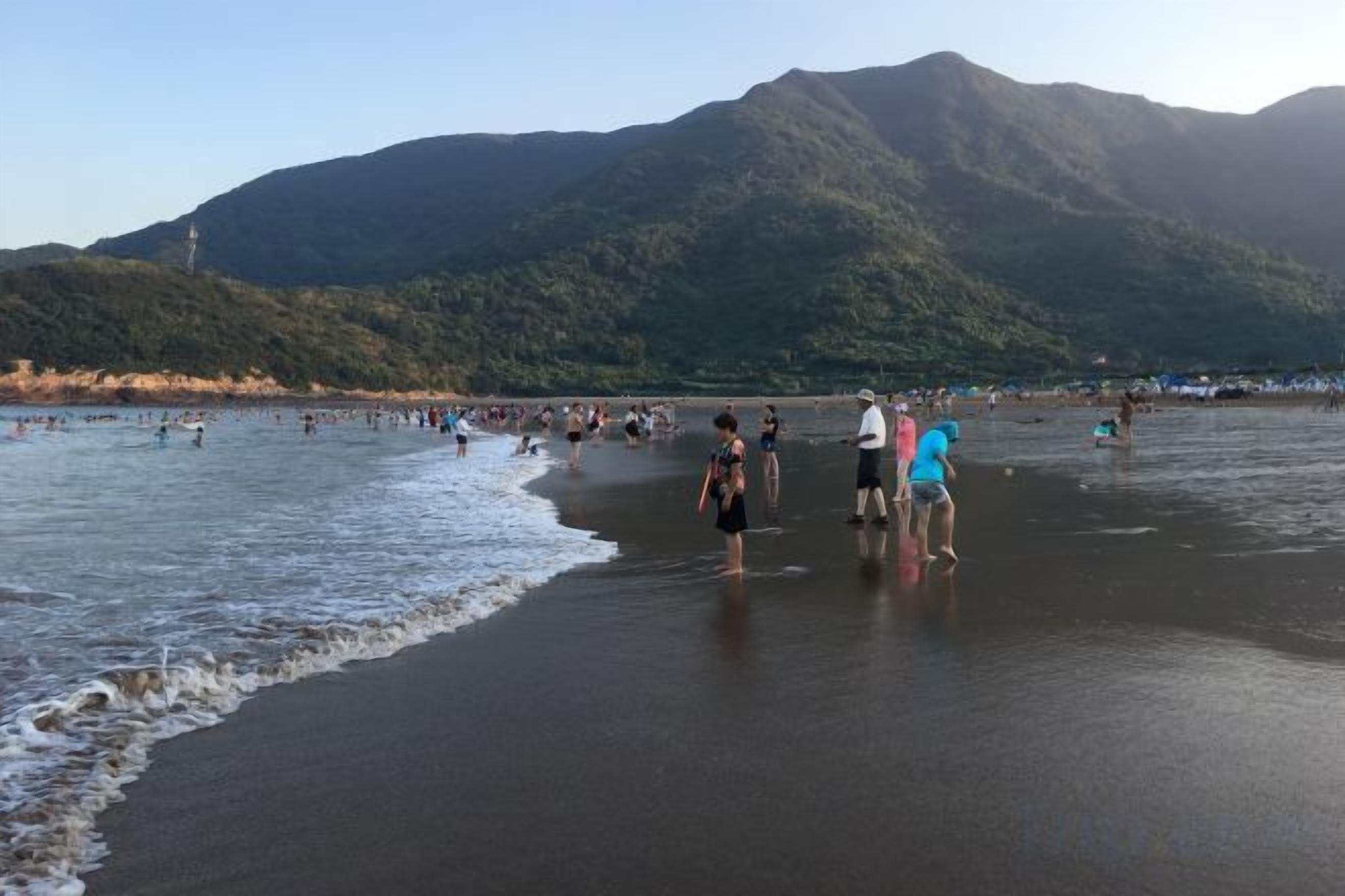 A beach with people standing in the water and on the sand, with mountains in the background.