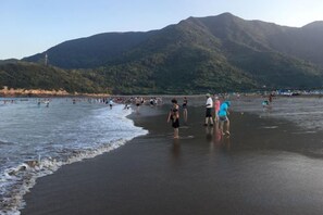 Una playa con gente de pie en el agua y en la arena, con montañas al fondo.