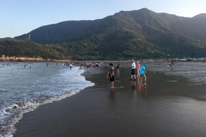 Una playa con gente de pie en el agua y en la arena, con montañas al fondo.