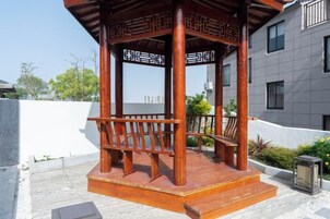 Un pabellón de madera con un banco, situado en un patio con un edificio moderno al fondo.