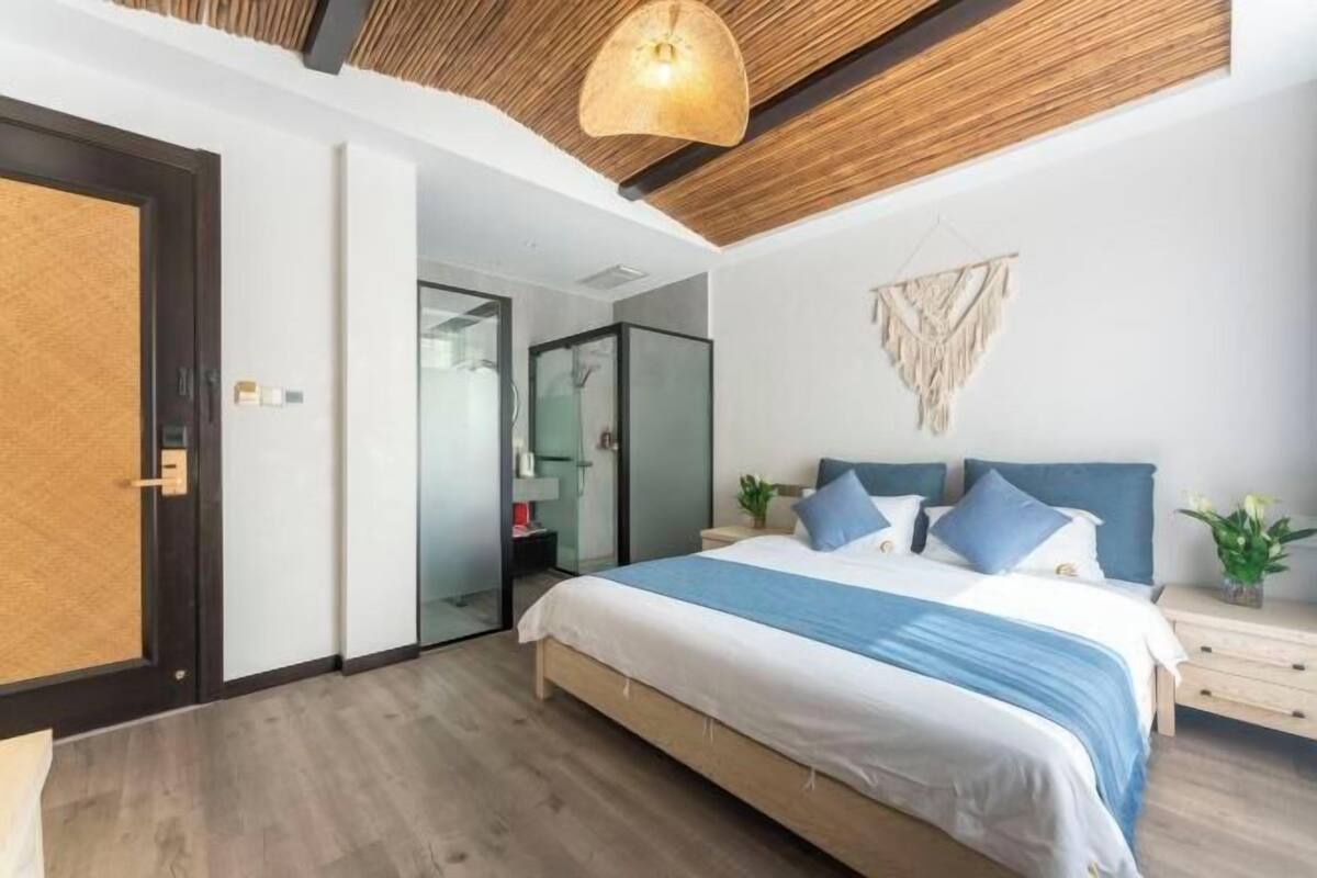 A modern bedroom with a wooden ceiling, a large bed with blue bedding, a bedside table, and a wall-mounted macrame piece.