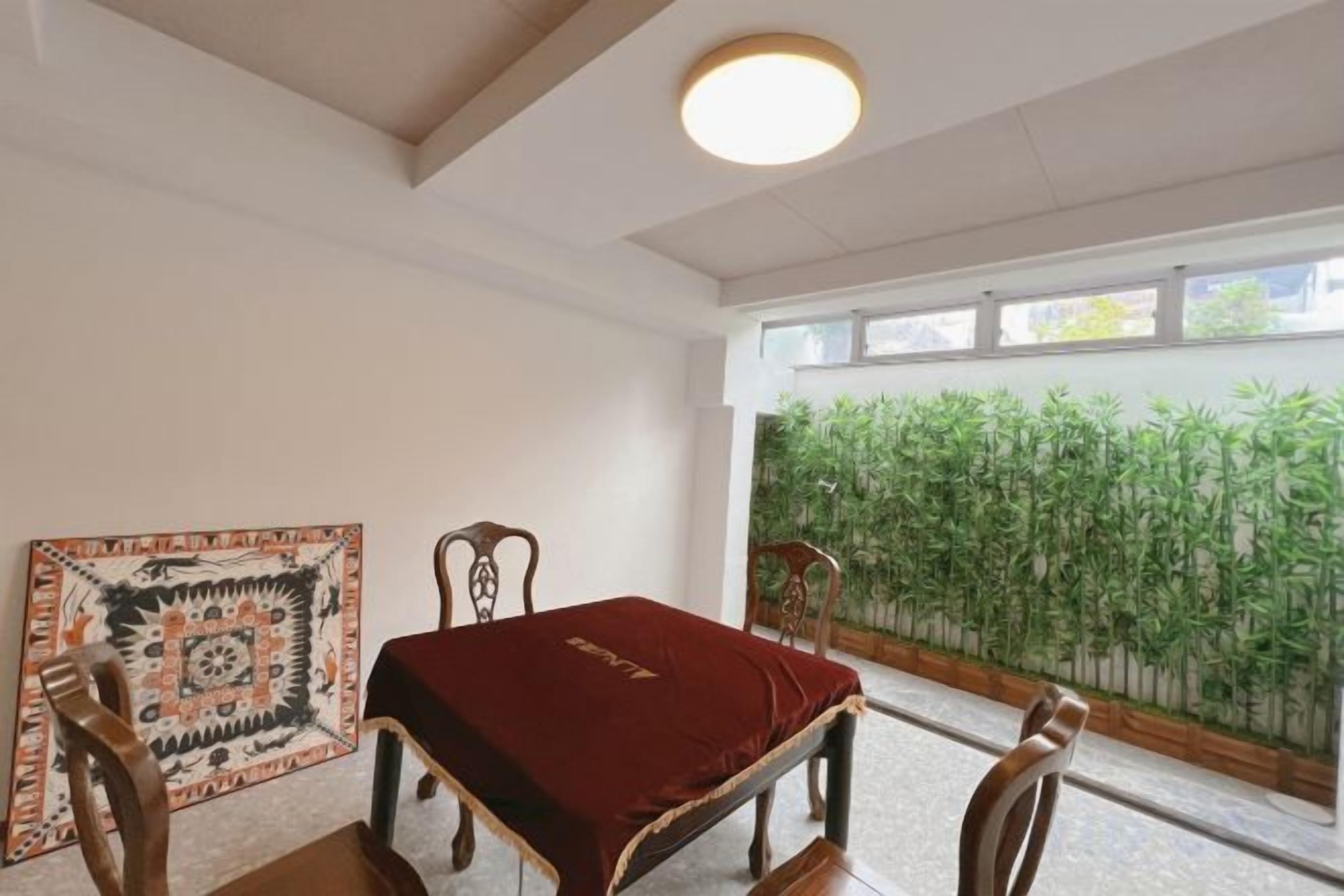 A room with a dining table, chairs, a patterned rug, and a view of bamboo plants through a window.