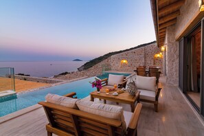 A poolside area with wooden furniture, a table with drinks, and a view of the sea.