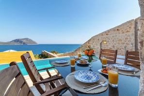 Una mesa de comedor con platos, cubiertos y vasos de zumo de naranja, con vistas a una piscina y el mar.