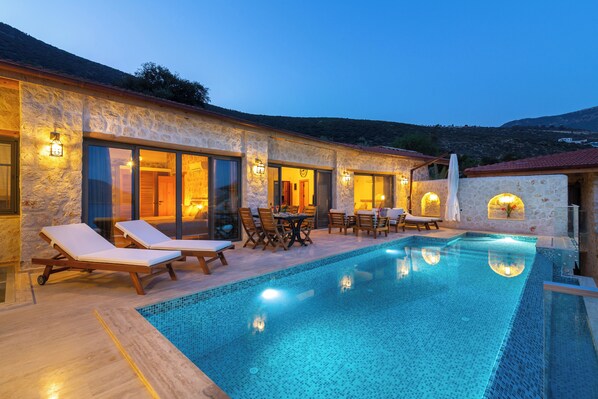 Un área junto a la piscina con sillones de descanso, una mesa de comedor y un edificio de piedra con amplias ventanas.
