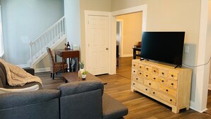 Una sala de estar con un sofá gris, un aparador de madera con cajones, un televisor y una escalera que lleva a otra habitación.