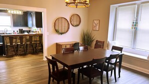 Un comedor con mesa y sillas de madera, una lámpara de araña y vista a la cocina de fondo.