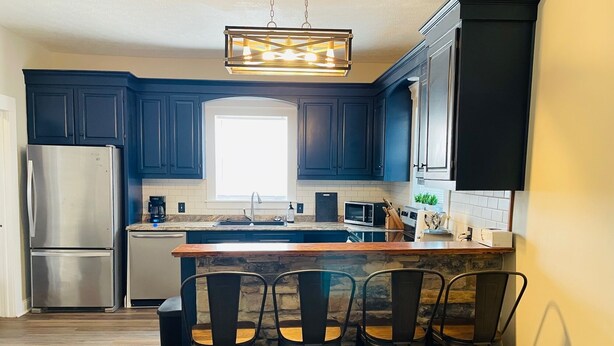 Una cocina moderna con armarios azul oscuro, un refrigerador de acero inoxidable, una isla integrada con taburetes altos y una lámpara colgante.