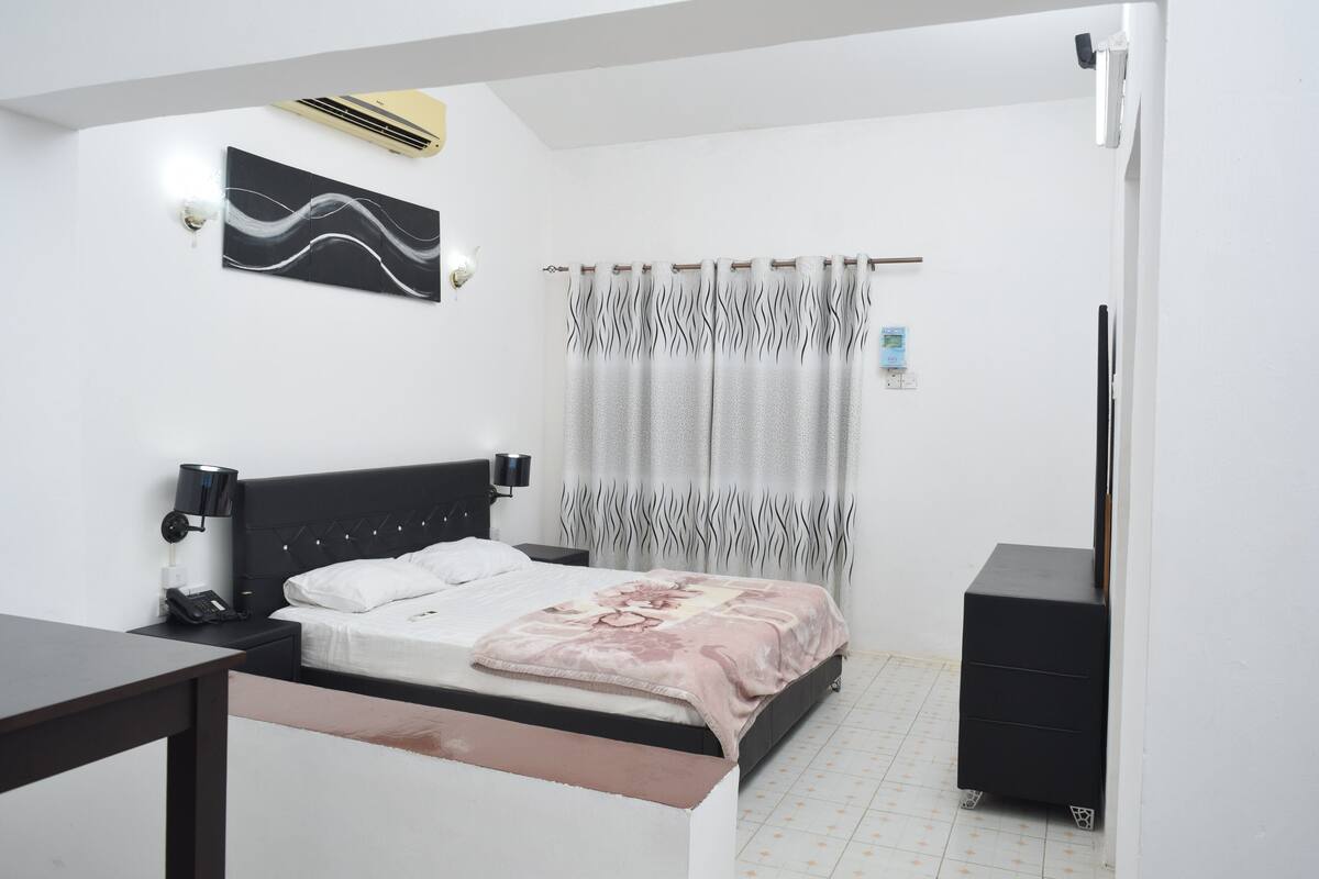 Une chambre à coucher comprenant un lit, une table de chevet, une lampe, un appareil de climatisation et un petit meuble.