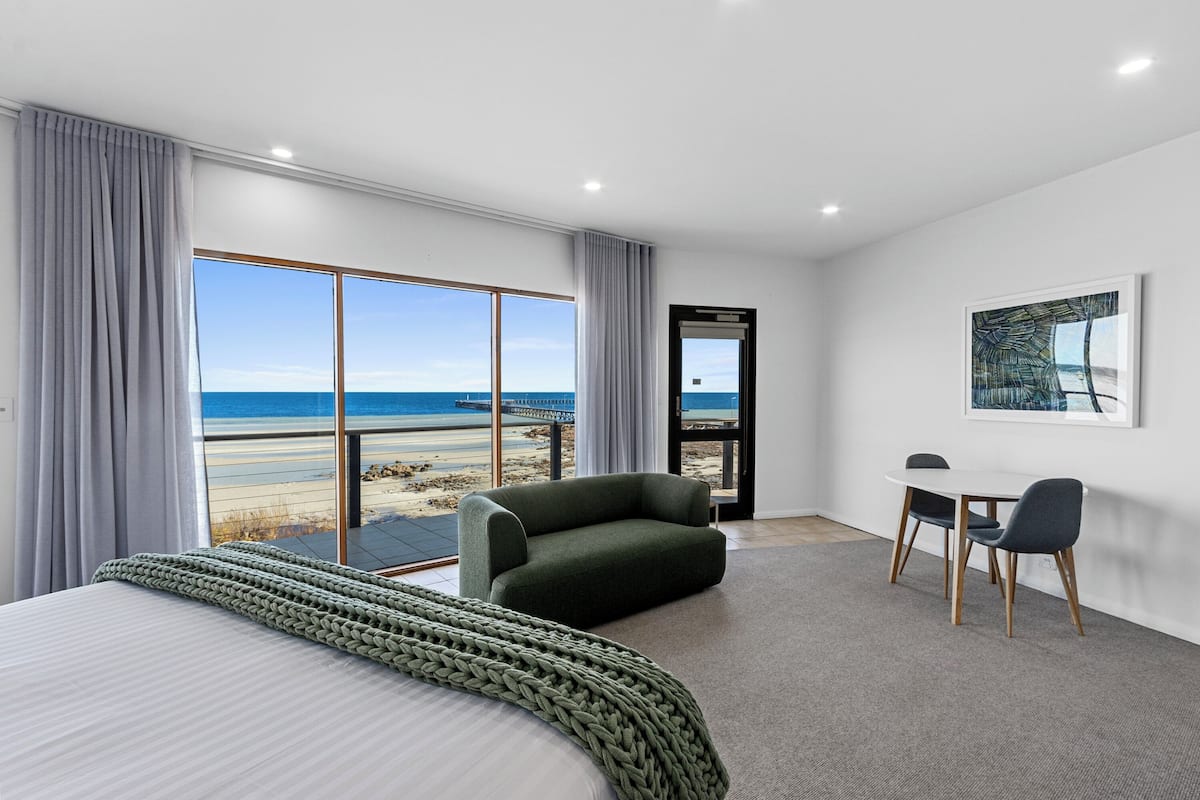 Un cuarto de hotel con una cama grande, un sillón verde, una mesita con dos sillas y vista a la playa a través de ventanas de piso a techo.
