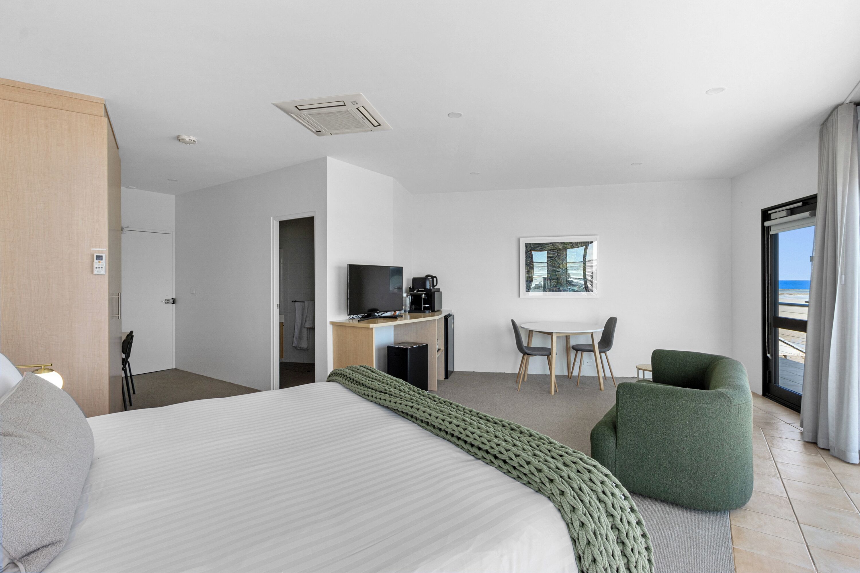 A hotel room with a large bed, a desk with a computer and coffee maker, a chair, and a view of the ocean.