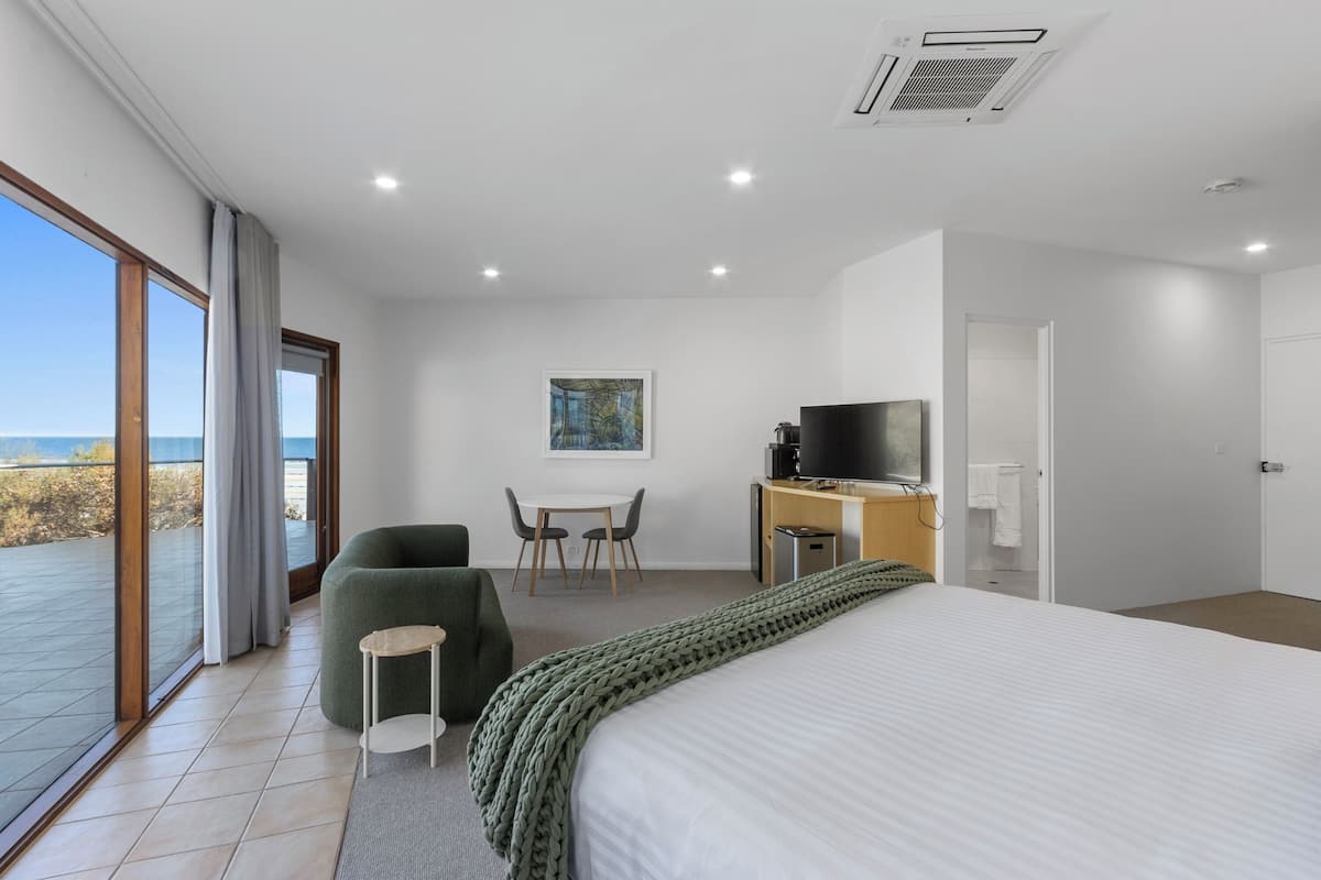 Una habitación de hotel moderna con una cama grande, un escritorio con televisor, un área de descanso con sillones y vista a la playa a través de la puerta de vidrio.