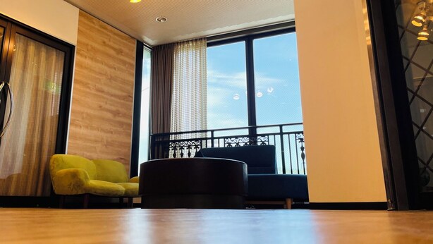 A modern living room with a wooden floor, a yellow armchair, a black sofa, and a large window with a view of tall buildings. - BUSINESS HOTEL KUDAMATSU (Kudamatsu)