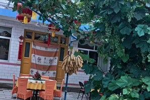 An outdoor dining area with a table set for two, a hanging bunch of bananas, and a building with a red and white striped awning. - Xinyuan Hostel (Jingtai)
