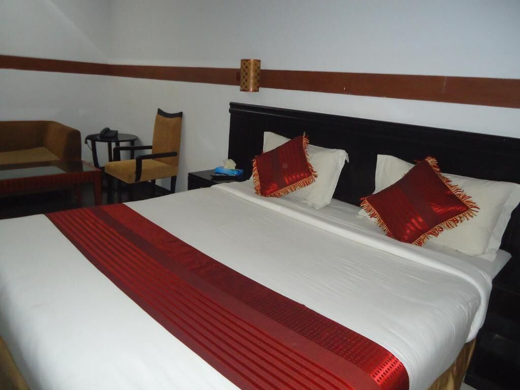 Una habitación de hotel con una cama grande, un cabecero de madera, dos sillas, una mesita y una mesita de noche con una lámpara.