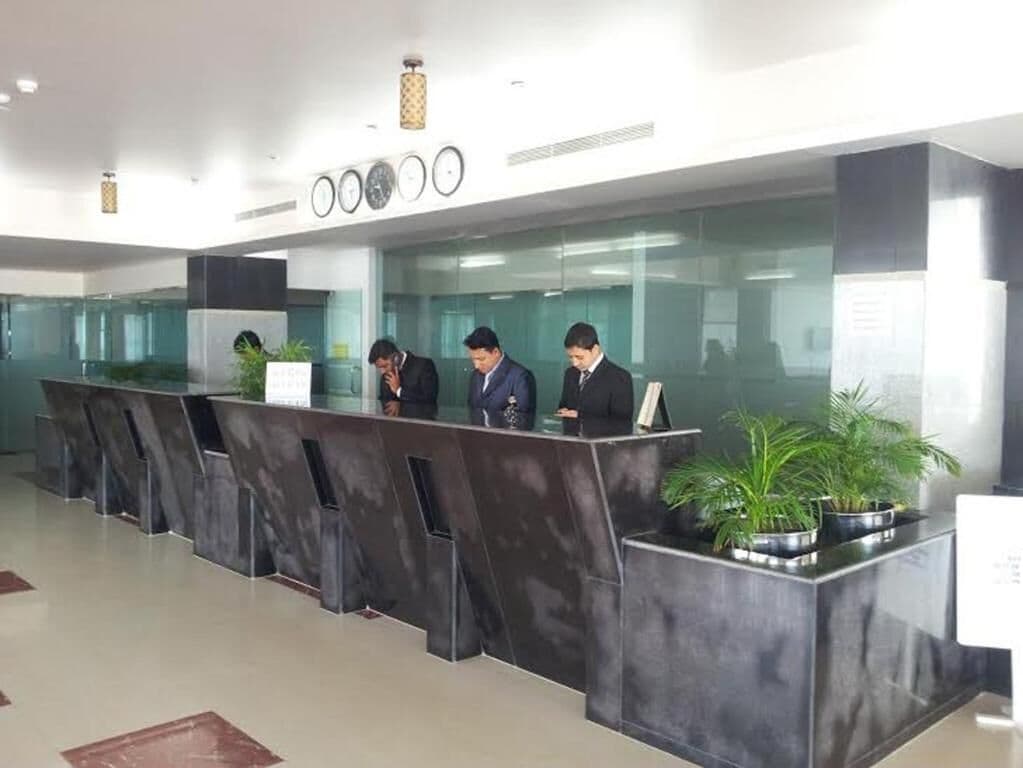 A reception area with a modern design, featuring a long reception desk with multiple counters, potted plants, and a clock on the wall.