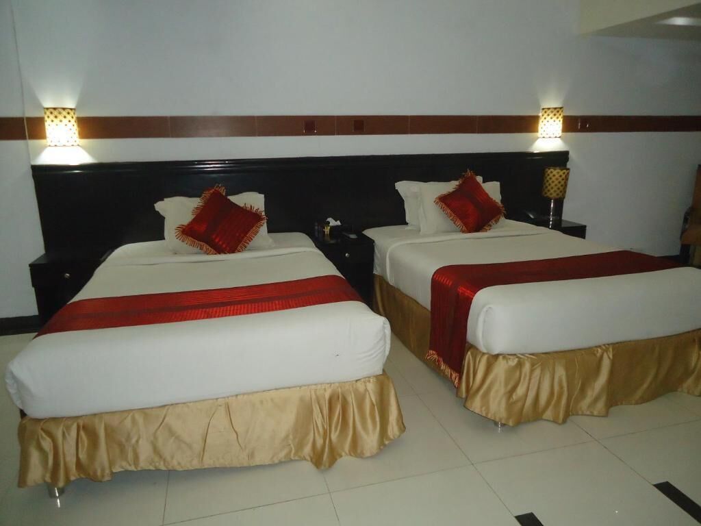 A hotel room with two single beds, each with a red and white bedspread, and a gold skirt. There are two bedside tables with lamps, and the room has a tiled floor.