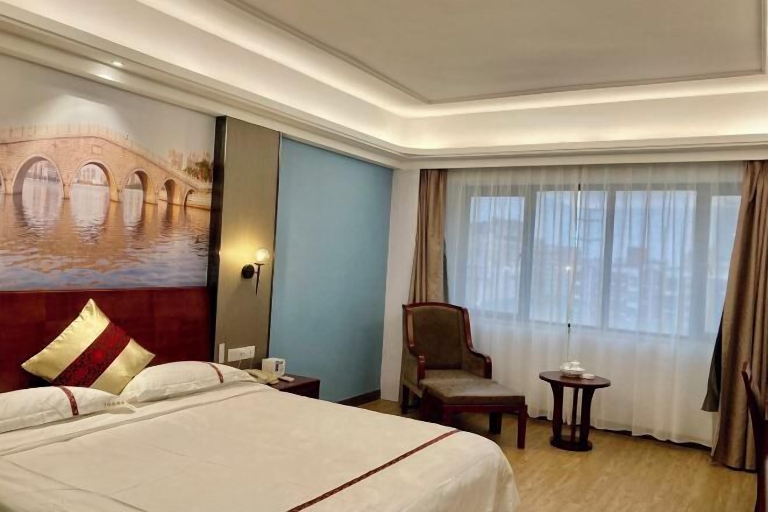 A hotel room with a large bed, a chair, a small table, a painting of a bridge, and a window with curtains.