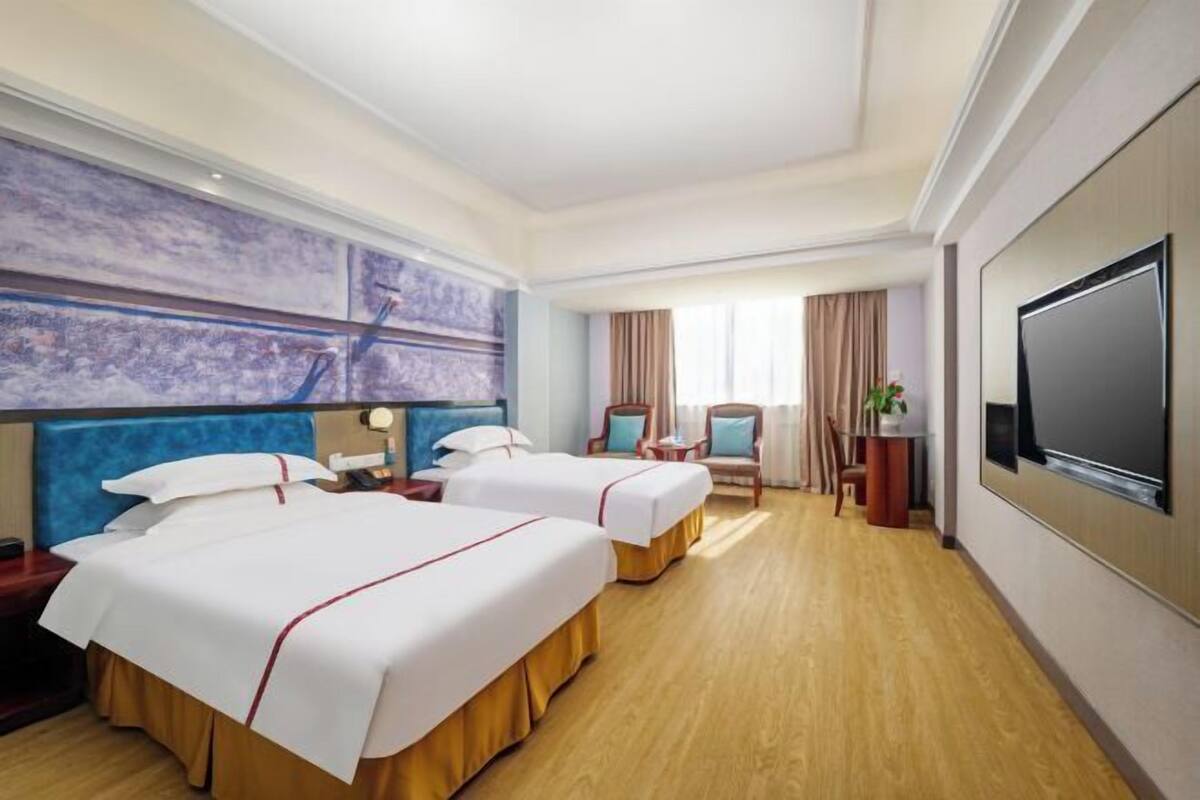 Une chambre d’hôtel avec deux lits, une télévision, un grand tableau et une fenêtre avec des rideaux.