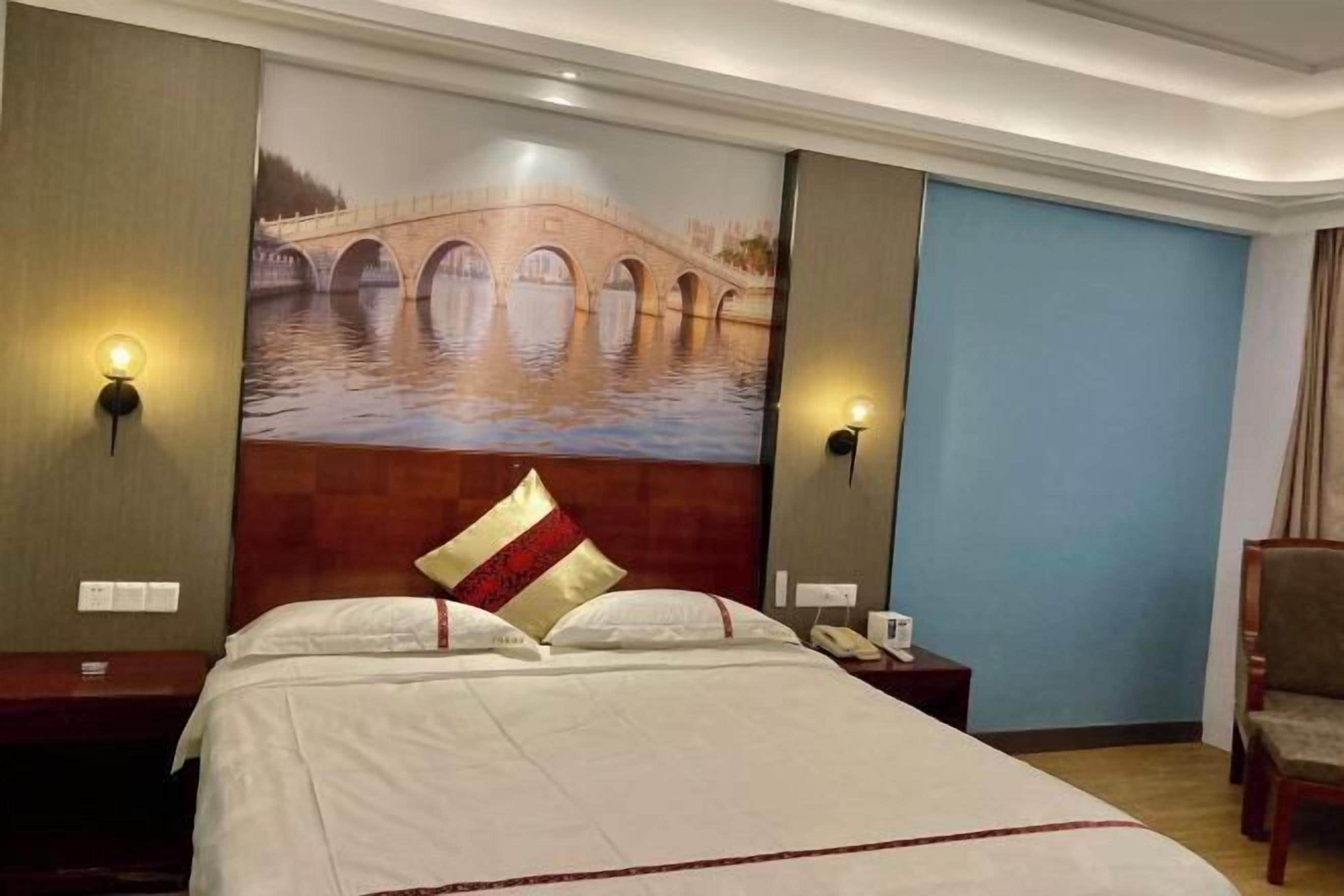 A hotel room with a bed, two bedside lamps, a nightstand, a chair, and a wall mural of a bridge over water.