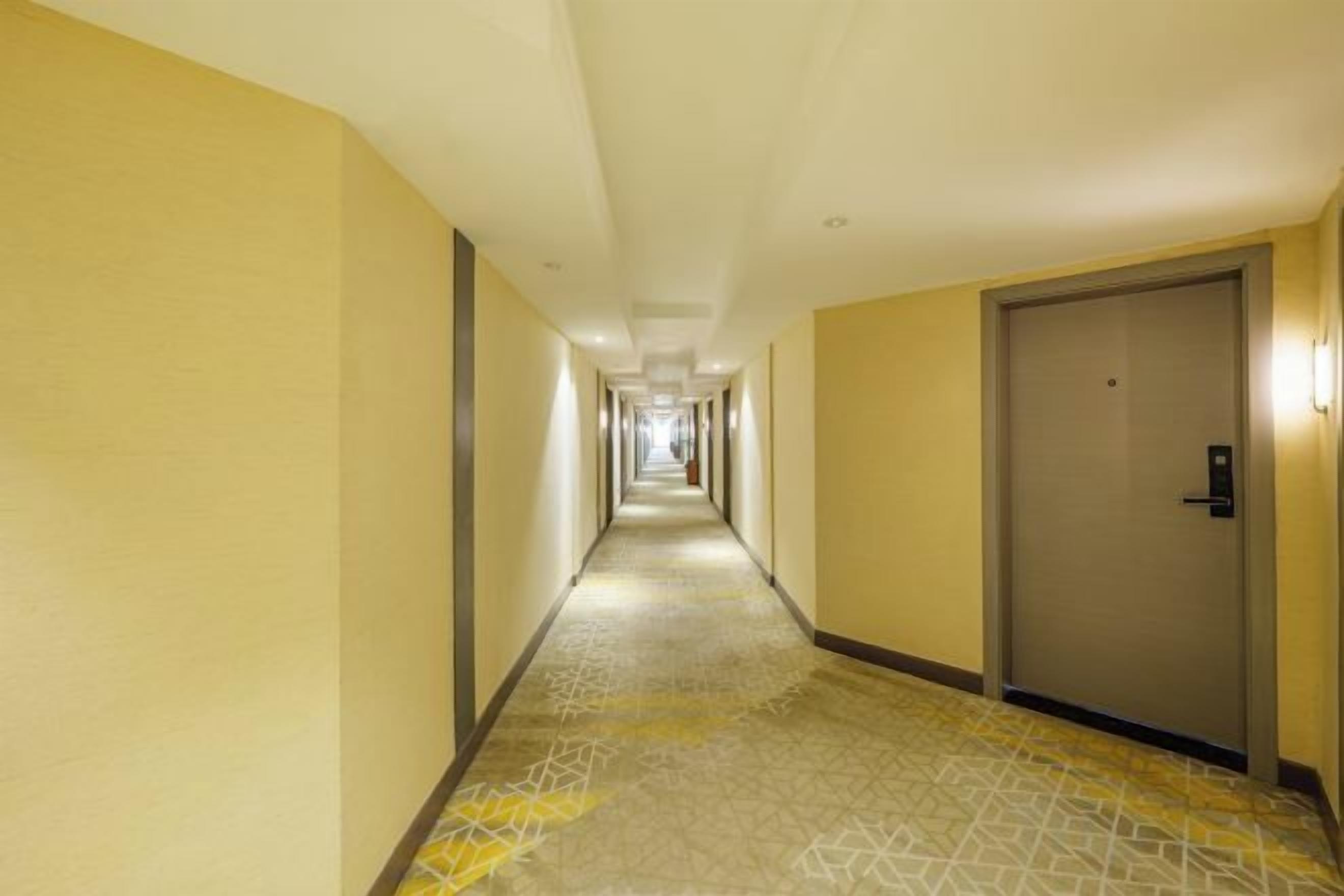A hotel corridor with a patterned carpet, a door on the right, and a light fixture on the wall.