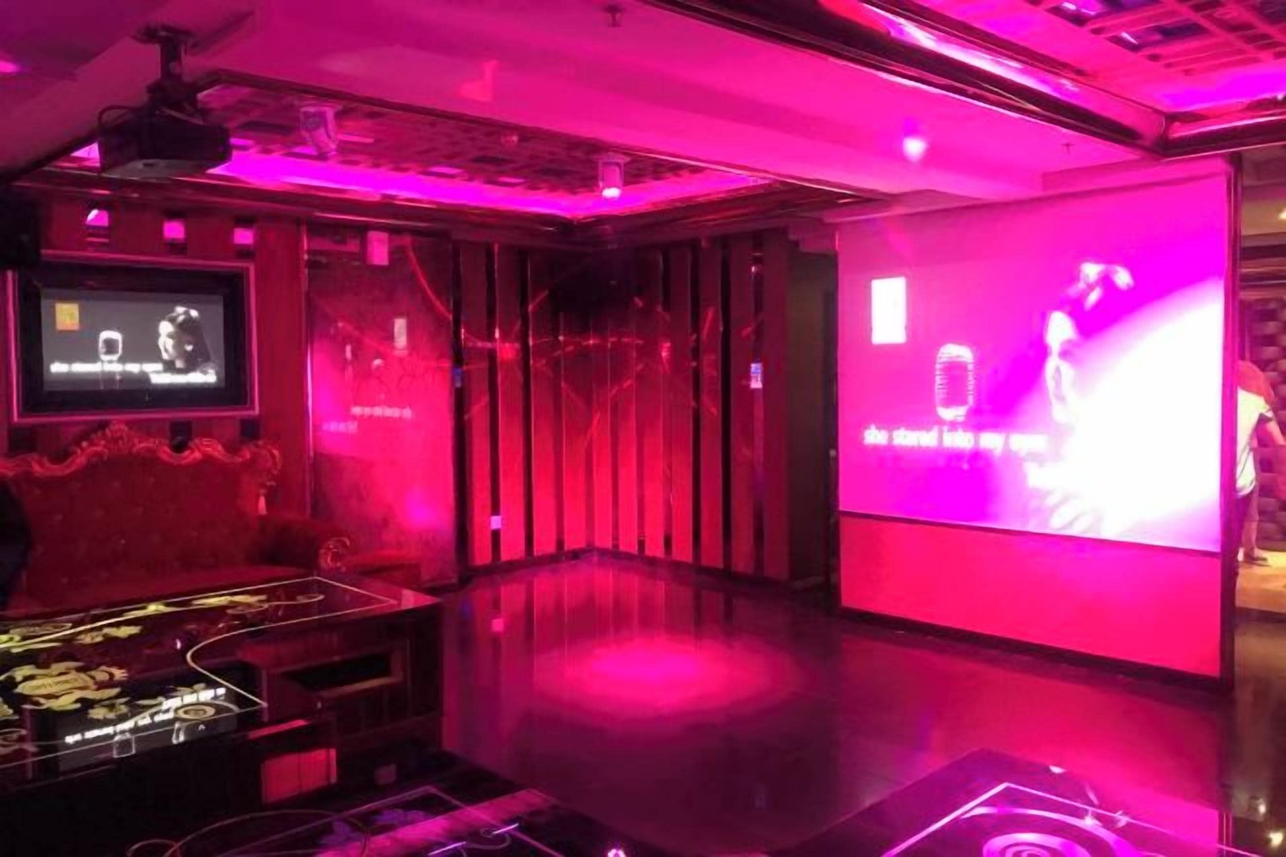 Una habitación con una pantalla grande mostrando una actuación, un televisor de pantalla plana y una zona de bar con encimera de vidrio.
