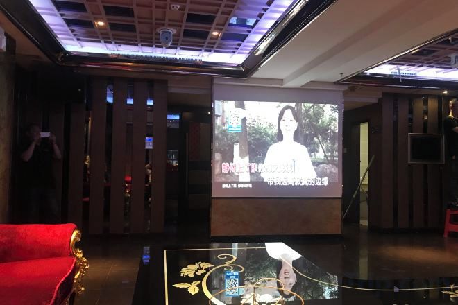 A lobby with a red sofa, a projection screen displaying a person, and a glass table with a decorative item.