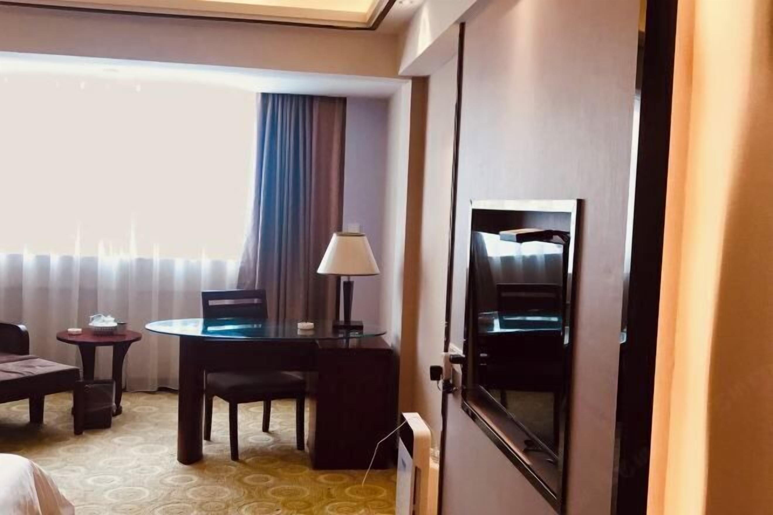A hotel room with a bed, a desk with a chair, a lamp, a TV, and a window with curtains.