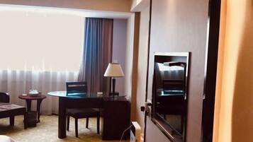 A hotel room with a bed, a desk with a chair, a lamp, a TV, and a window with curtains.