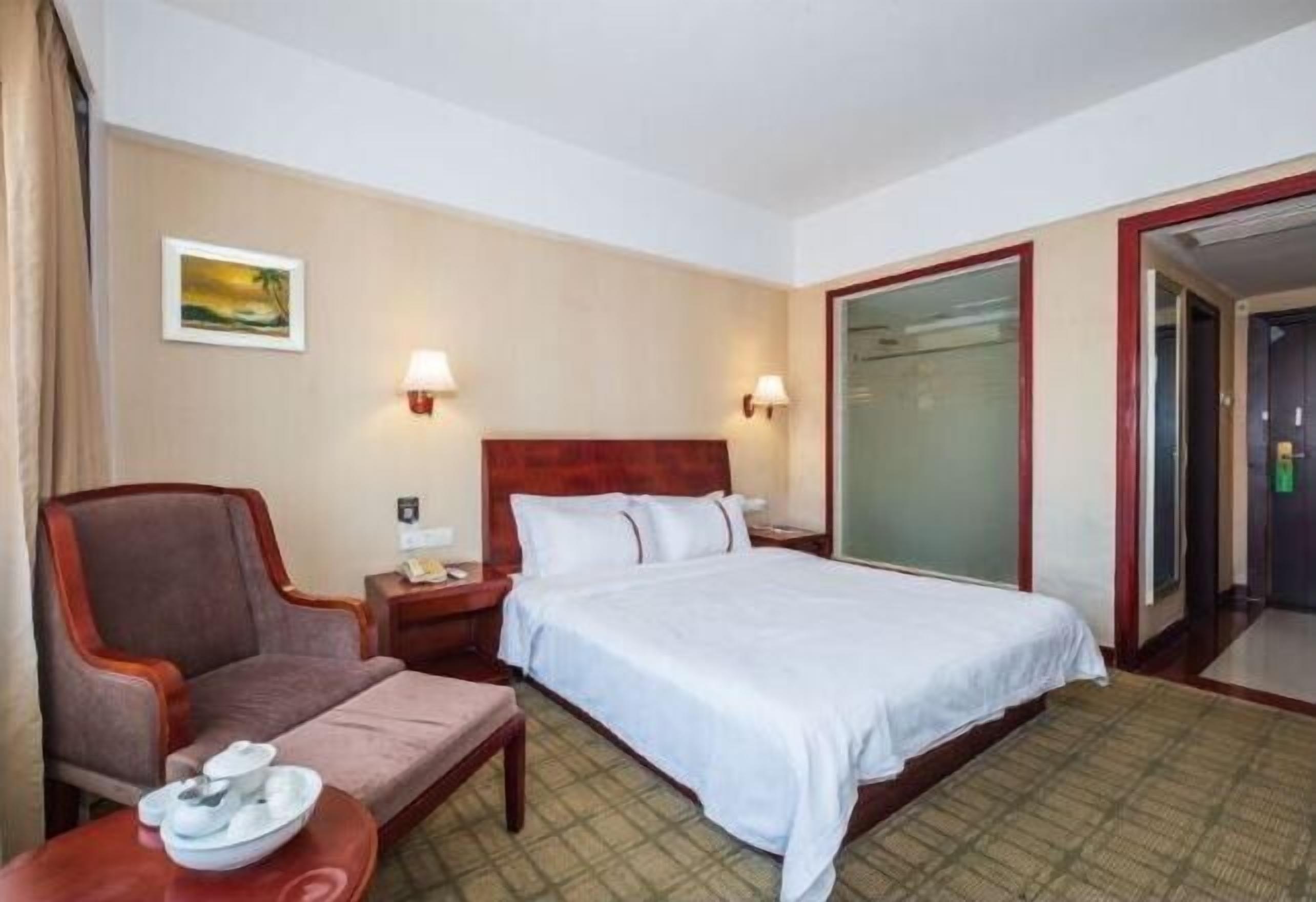 Quarto de hotel com uma cama, uma cadeira, uma mesa de cabeceira e uma luminária fixada na parede.