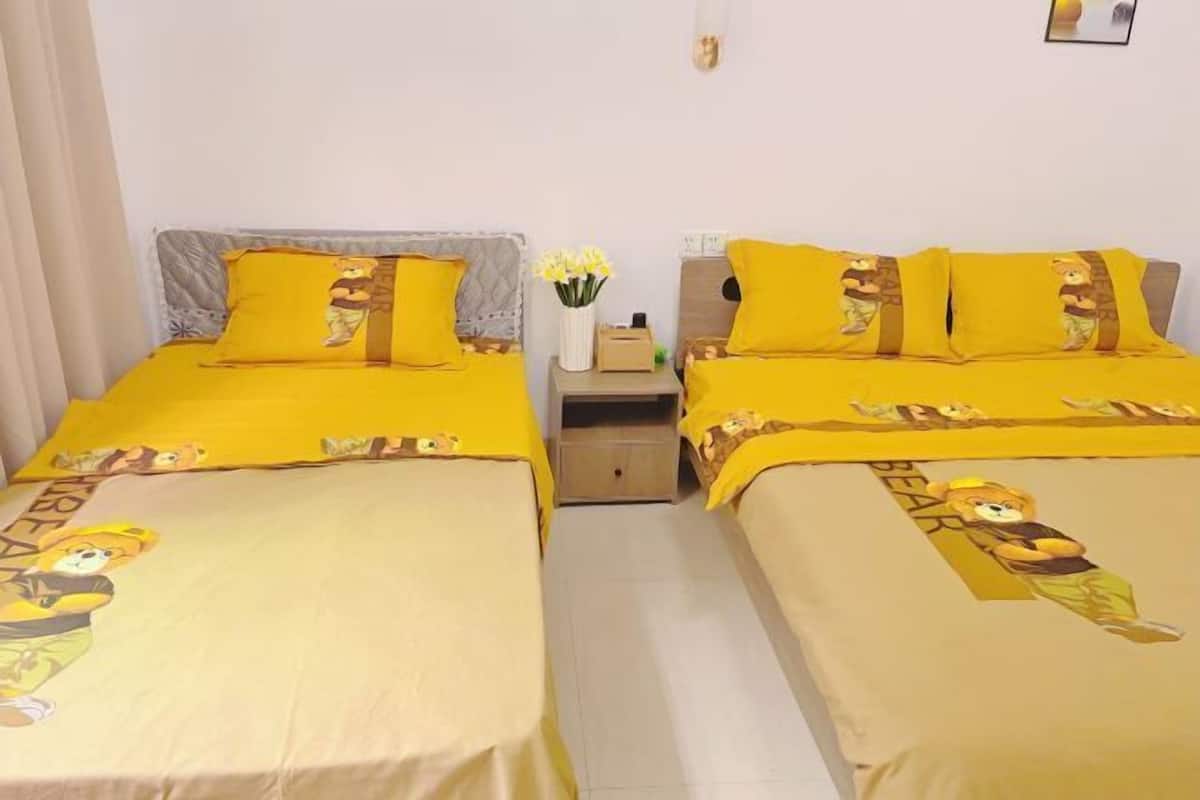 A room with two beds, each with yellow bedding and cartoon character prints, a small bedside table with a vase of flowers, and a wall-mounted light fixture.
