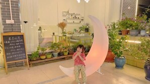 Un nen dempeus en una habitació amb una decoració de creu lunar, plantes en testos i una pissarra amb text.