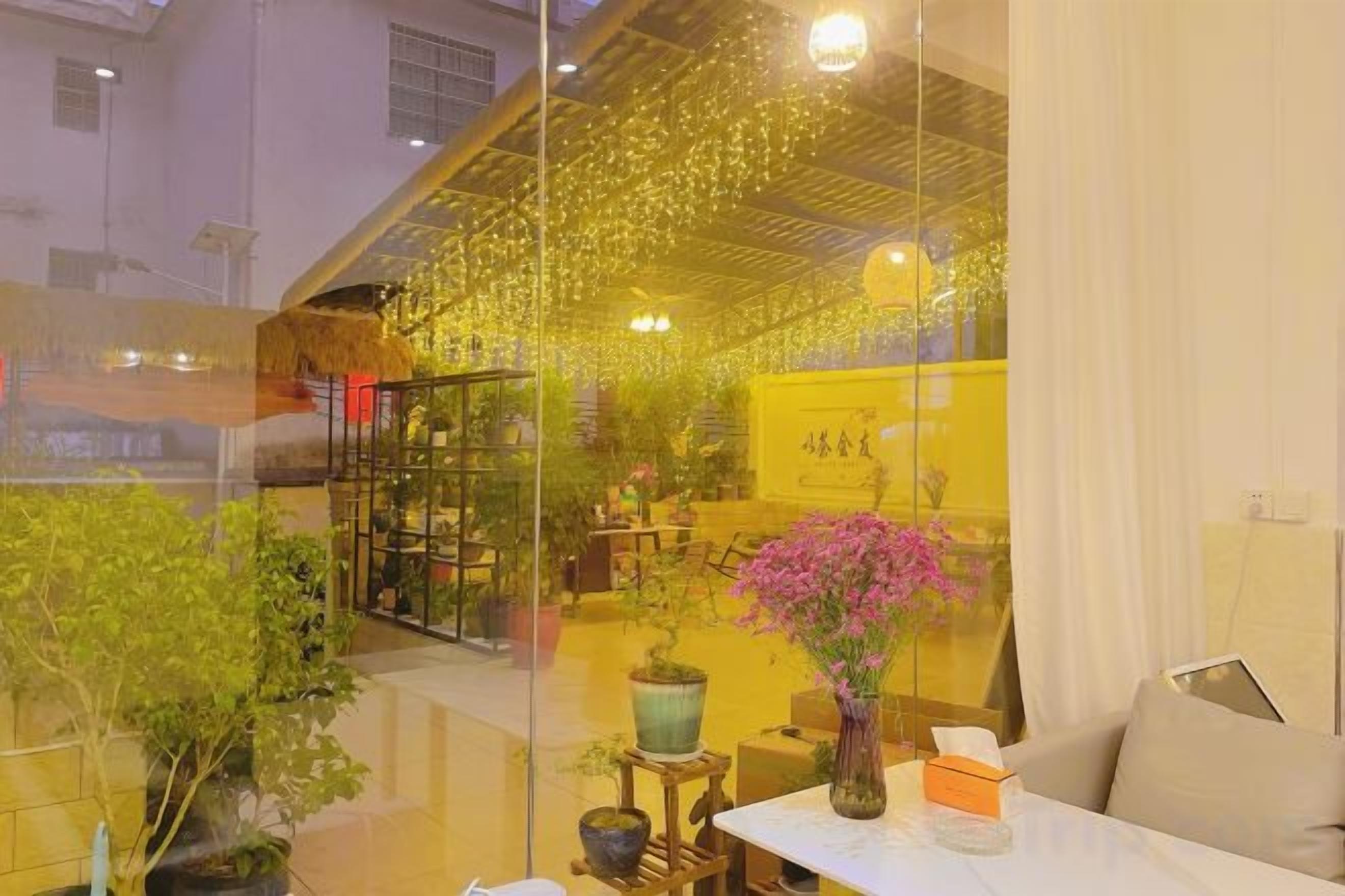 A room with a glass partition, a table with a vase, and a chair. There are potted plants and a yellow sign in the background.