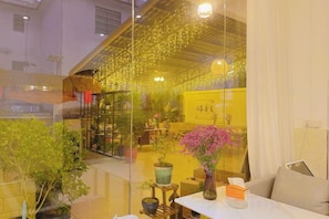 A room with a glass partition, a table with a vase, and a chair. There are potted plants and a yellow sign in the background.