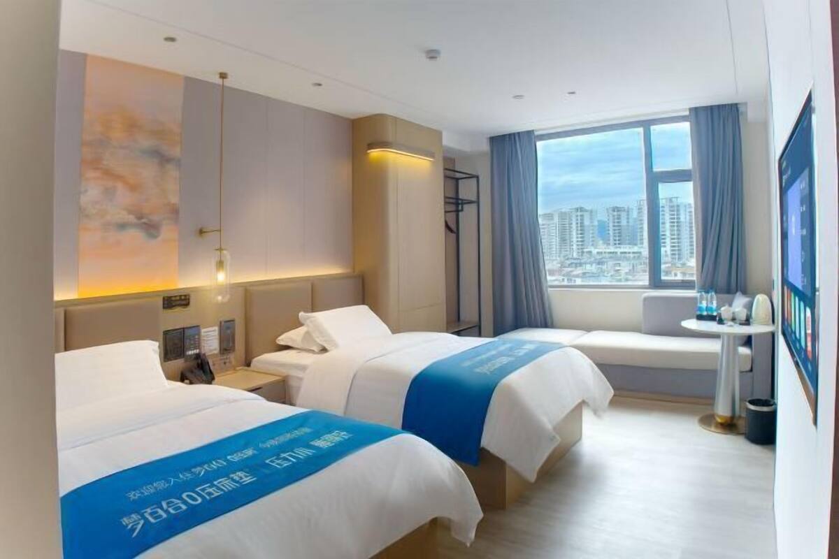 A hotel room with two beds, a desk, a chair, a window with a city view, and a painting on the wall.