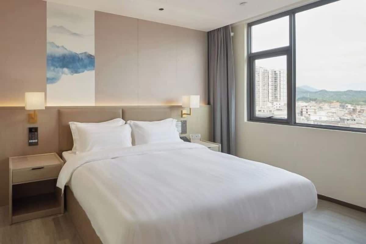 A hotel room with a bed, bedside tables, a nightstand, a wall-mounted lamp, and a window with a city view.
