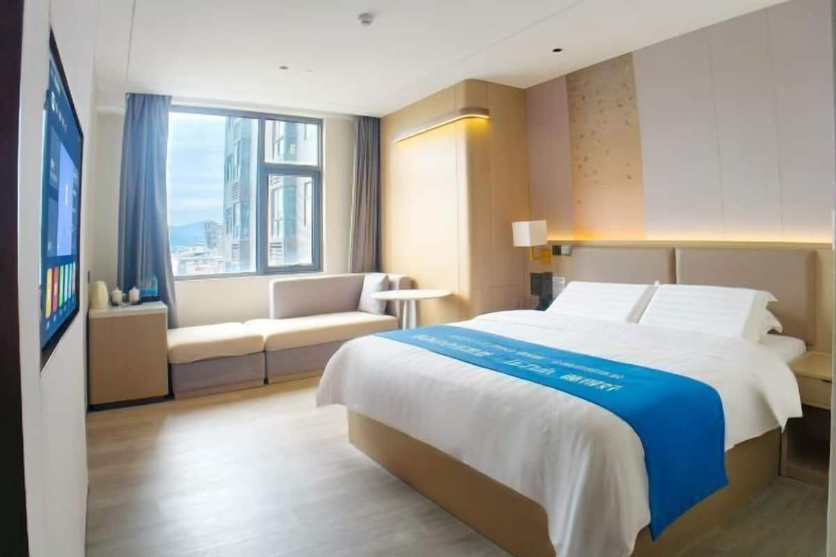 A hotel room with a large bed, a seating area, a window with a city view, and a wall-mounted TV.